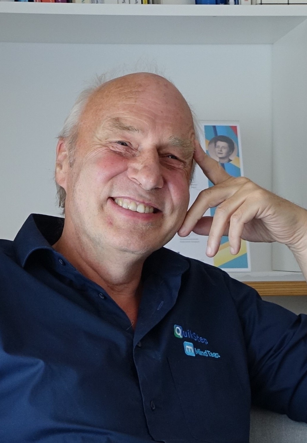 (Selbstbeschreibung) Ein älterer Mann, Stefan Wilke Geschäftsführer der QuikStep GmbH, sitzt entspannt vor einem hellen Regal und lächelt freundlich in die Kamera. Er trägt ein dunkelblaues Hemd mit den Logos QuikStep und MindTags auf der Brust. Sein Kopf ist leicht zur Seite geneigt, während er mit der Hand den Kopf abstützt – eine offene, nachdenkliche und zugleich sehr sympathische Haltung. Sein Haar ist kurz und graues Haar und er zeigt ein warmes, herzliches Lächeln. Im Hintergrund steht ein weißes Bücherregal mit einigen Büchern und einem kleinen gerahmten Bild mit bunten Farben. Die Atmosphäre wirkt ruhig, persönlich und vertrauensvoll.